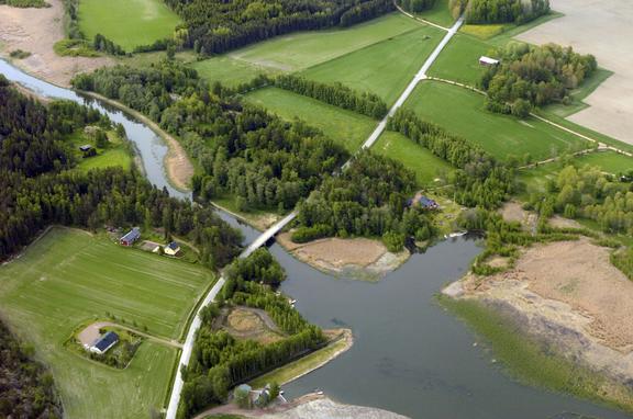 An aerial view of a rural landscape where a winding river or narrow waterway flows through fields and wooded areas. A road crosses the water via a bridge. The image also shows scattered houses and farm buildings, extensive green farmland, and reed-covered shoreline areas along the water.